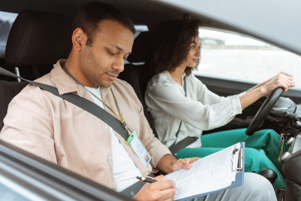 Arabic Instructor Taking Notes Teaching Novice Driver Woman Driving Car Driving Test. Arabic Instructor Man Taking Notes In Lesson Plan Teaching Novice Driver Woman Sitting In Car. Coach Mastering And Evaluating Learner's Practical Skills. Selective Focus, Side View